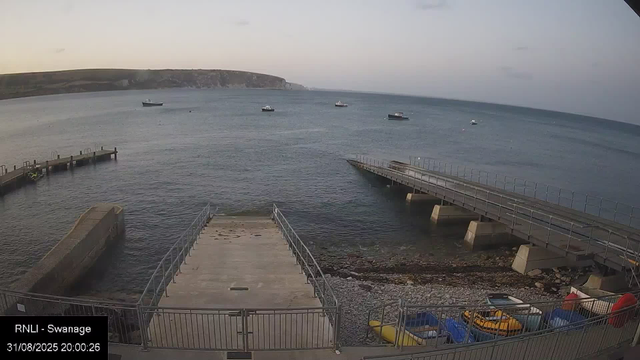 A view of a coastal area at sunset with a calm sea containing several boats anchored in the distance. In the foreground, there are two concrete piers or jetties extending into the water, one on the left and one on the right, with railings along their edges. At the bottom of the image, there is a fenced area with small boats, some yellow and blue, as well as orange life rings. The sky is partly cloudy, and a distant landmass stretches across the horizon.
