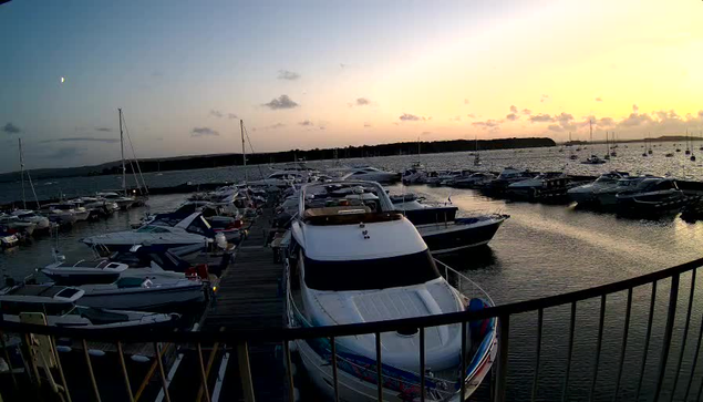 A marina filled with various boats and yachts docked at a wooden pier during sunset, with the sky painted in soft shades of orange and pink and calm water reflecting the colors.