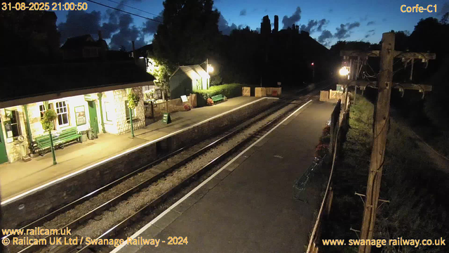 A small train station platform is illuminated by yellowish lights, with a building on the left side that has green window frames, doors, and benches. The platform is empty with no passengers. The railway tracks run parallel to the platform, extending into the distance. On the right side, there are wooden utility poles with wires and some flower arrangements in pots. The sky is dark with a few clouds, indicating that it is early evening or night.