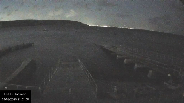 A dark, low-light image showing a large, empty outdoor area near buildings. The sky is dark, indicating nighttime, with some clouds visible. In the lower left corner, there is a timestamp reading "31/08/2025 21:01:06" and text indicating "RNLI - Swansea."