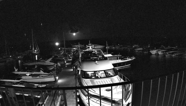 A black-and-white photo taken from a webcam shows a marina at night with multiple boats docked. In the foreground, there is a curved railing, and behind it, a walkway with several boats moored on either side. The boats vary in size, with some having tall masts and others being motor yachts. Some boats have visible cabin windows reflecting light, and the marina appears calm and quiet under the dark sky, with minimal lighting illuminating the scene.