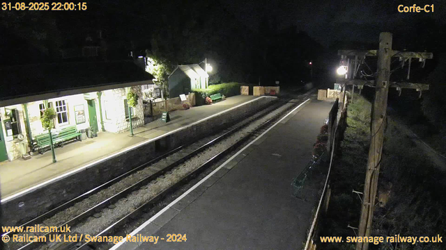 A small train station platform at night, illuminated by a few lights. On the left side, there is a building with green door and window frames, decorated with hanging plants and a bench outside. A sign indicates "Way Out." In the middle, the train tracks run parallel to the platform, which is empty. On the right side, there are wooden utility poles and fencing, with some flower arrangements and a few benches along the platform. The sky is dark, and the surrounding area is not well-lit.