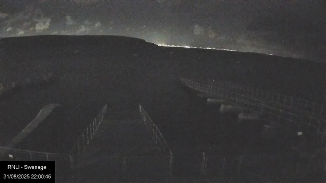 A dark, nighttime view from a webcam showing a field with fencing in the foreground. In the background, faint lights are visible along the horizon, possibly from distant buildings or houses. The sky is mostly dark with some clouds, and the scene appears to be quiet and empty. The timestamp reads "31/08/2025 22:00:46," and there is a label indicating this is from the RNLI in Swanage.