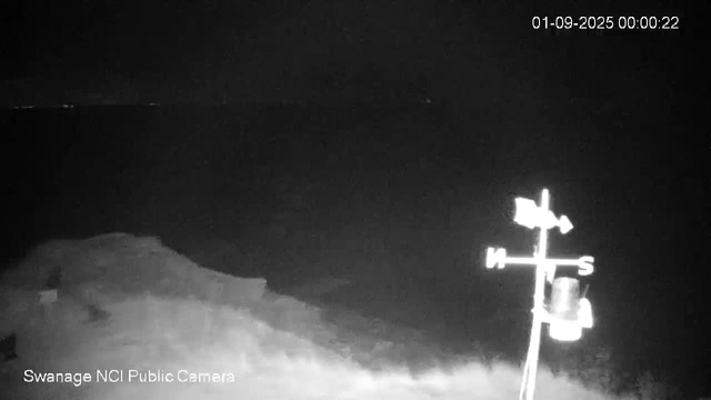 A nighttime scene showing an outdoor landscape with a weather vane in the foreground. The weather vane has directional markers labeled "N," "S," "E," and "W," and is mounted on a pole with a small cylindrical object attached below it. The background is dark with no visible structures or objects illuminated. The timestamp on the top right corner reads "01-09-2025 00:00:22," and text at the bottom indicates the image is from "Swanage NCI Public Camera."