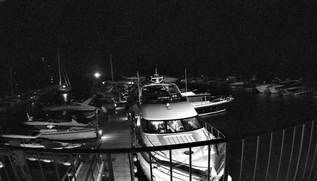 A nighttime scene of a marina filled with various docked boats and yachts. The boats are aligned along a pier, illuminated by some lights, with several sailboats and motorboats visible in the background. The sky is dark, and the water appears calm. A railing is visible in the foreground, suggesting the photo was taken from a nearby balcony or walkway.