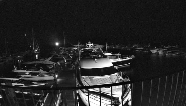 A dark nighttime scene at a marina with multiple boats docked. In the foreground, a large yacht is visible behind a metal railing, with other boats moored in the background. The water is calm and the scene is dimly lit, possibly by a single exterior light or moonlight.