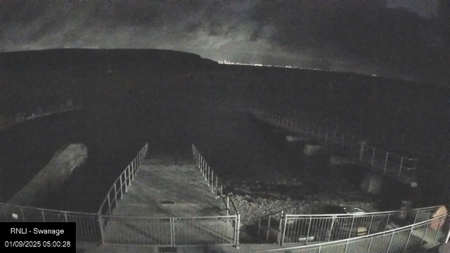 A nighttime view of a construction site or fenced area with a large, curved, black structure, possibly a bridge or an overpass. The scene is dark, with minimal lighting, and the area is surrounded by a metal fence. The sky is cloudy or overcast, and the ground appears uneven with some construction materials or debris.