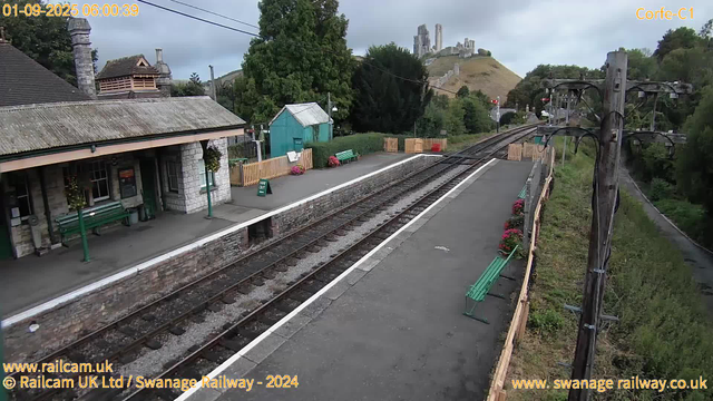 A small train station platform with a stone building on the left, featuring green benches and hanging flower baskets. There are wooden fences, a green bench, and a small teal shed behind the platform. The railway tracks run through the center, with a small sign that reads "WAY OUT" on the platform. In the background, there are green trees and a hill with ruins of a castle on top. To the right, there are electrical poles with wires, and a narrow path or road running alongside the railway, bordered by greenery. The sky is cloudy and overcast. The image has a timestamp in the top left corner showing "01-09-2025 06:00:39."