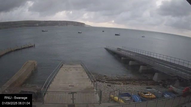 A cloudy sky over a calm body of water with several boats floating in the distance. In the foreground, there are two piers extending into the water, one on the left with a concrete surface and railing, and the other on the right with a metal walkway. At the bottom of the image, there is a fenced area with small colorful boats and kayaks. The time and date in the corner indicate early morning on September 1, 2025, at 6:01 am, with the location labeled as RNLI - Swanage.