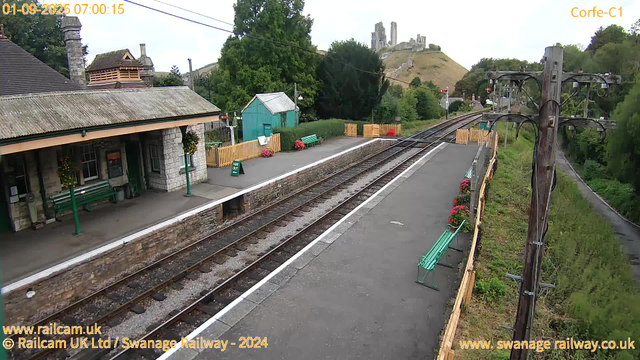 A small train station platform with a stone building on the left, decorated with hanging flower baskets. There are green benches, a small turquoise shed, and colorful flowers along the fence. Parallel train tracks run through the station, with a winding railway line continuing into the distance. A wooden utility pole with electrical wires stands on the right side next to a grassy embankment. In the background, a hill with a ruined castle sits atop, surrounded by trees. The sky is overcast with a soft light, and the scene appears calm and quiet early in the morning.
