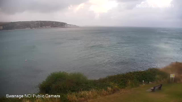 A view of a coastline with a body of water in the foreground and a cliff or headland in the distance on the left. The sky is mostly cloudy with some sunlight breaking through. There is greenery and a bench in the lower right corner, along with some small signs. The scene appears calm and natural.