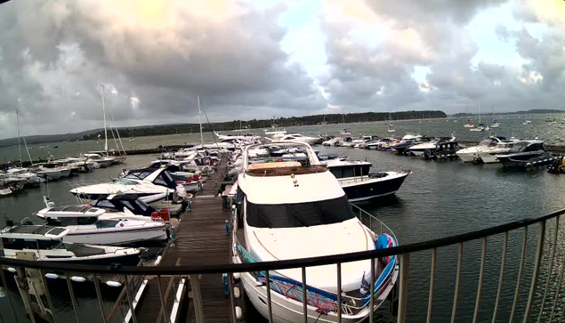 A marina filled with numerous boats and yachts docked along wooden piers. The sky is partly cloudy with dark clouds and patches of sunlight, and in the background, there is a distant shoreline with some trees.