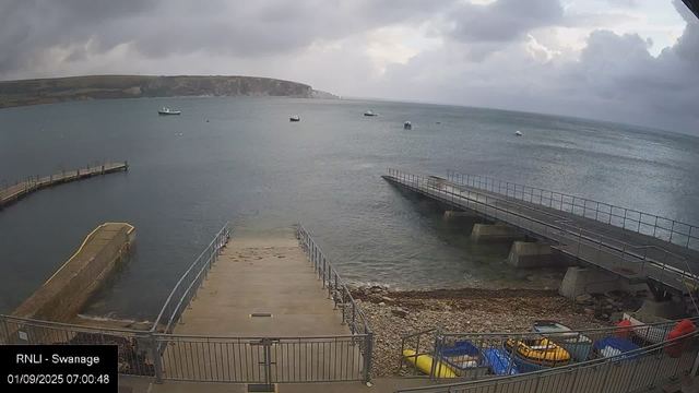 A view of a harbor with a concrete pier extending into the water, surrounded by metal railings. Several small boats are anchored on the calm sea. In the background, there is a low, rocky shoreline with some cliffs under a cloudy sky. The scene appears to be in the early morning, as indicated by the timestamp "01/09/2025 07:00:48."