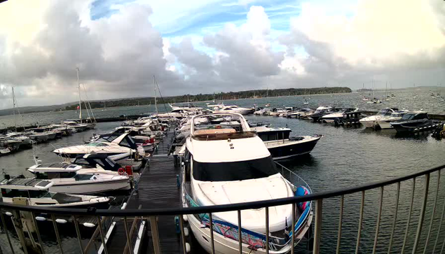 A marina filled with numerous boats and yachts docked along wooden piers, with some boats featuring covered decks. The water is calm, and there are sailboats visible in the background besides the larger yachts. The sky above shows a mix of clouds and patches of blue, with a distant shoreline visible across the water.