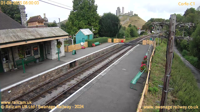 A small train station platform with a stone building on the left, decorated with hanging flower baskets. There are green benches on the platform, and a small turquoise shed at the far end. Bright yellow fencing surrounds some flower beds with pink flowers. There are power lines overhead and a wooden pole on the right side of the image. In the background, a hill with ancient castle ruins is visible. The sky is cloudy and overcast. The timestamp indicates the date and time as 01-09-2025 at 08:00:03.