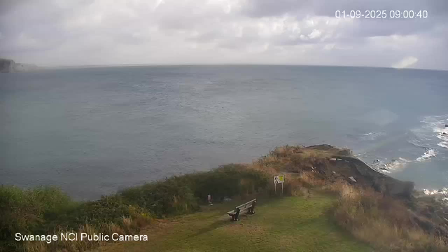 A view of a coastal landscape showing the ocean under a cloudy sky. In the foreground, there is a grassy area with a bench, a person sitting on the bench, and a signpost. The coastline features rocky cliffs with waves crashing against them. The timestamp in the upper right corner reads "01-09-2025 09:00:40."