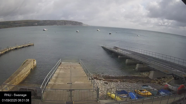 A view of a harbor with a concrete pier extending into the water, surrounded by a metal railing. To the left, there is a small jetty made of concrete with metal handrails. On the right, a longer, slightly elevated pier with metal railings extends into the water. The shoreline has rocks and pebbles, with small boats and kayaks stored close to the fence at the bottom of the image. Several boats are floating on the water in the distance, and the sky is cloudy with patches of light breaking through. A large landmass with cliffs is visible on the horizon.