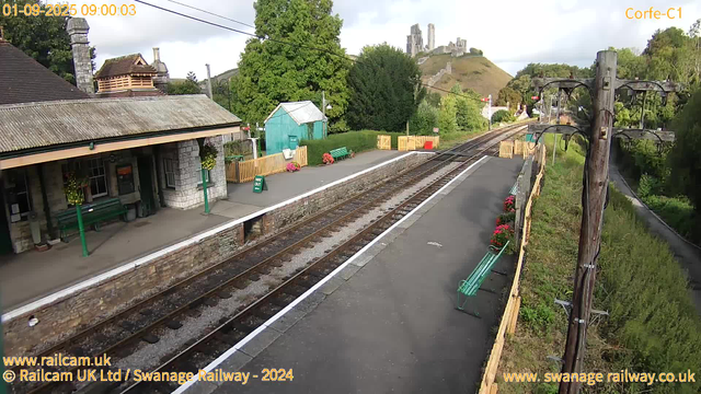 A small countryside train station platform with a stone building on the left, decorated with hanging flower baskets. There are green benches, a small turquoise shed, and yellow barriers along the platform. Overhead, electrical wires are supported by a tall wooden pole on the right side, with a few flowers in pots near the edge of the platform. In the background, green trees and a hill with a castle ruin on top are visible, under a partly cloudy sky.