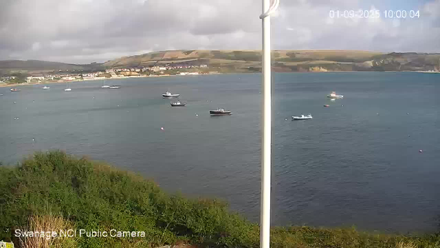 A view of a harbor with several boats floating on calm water, a cloudy sky overhead, and rolling hills in the background. There is a white pole in the center of the image. Green foliage is visible in the foreground. The timestamp in the upper right corner shows the date and time as January 9, 2025, at 10:00 AM. The bottom left corner indicates the image is from the Swannage NCI Public Camera.