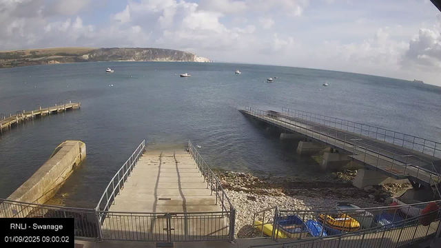 A view of a seaside area with a concrete pier extending into the water, surrounded by railings. There are a few small boats floating on the water and some parked on the shore. The background shows a distant cliff and a partly cloudy sky. The timestamp indicates the image was taken on September 1, 2025, at 9:00 AM.