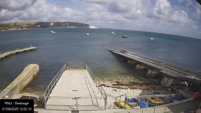 A view of a seaside harbor with several boats anchored in the water. There are two piers extending into the sea, one on the left and one on the right. The foreground shows a concrete platform with metal railings, and some small boats or kayaks are stored at the bottom. In the background, there are cliffs and a partly cloudy sky. The timestamp on the image reads "01/09/2025 12:00:10" and it is labeled "RNLI - Swanage."