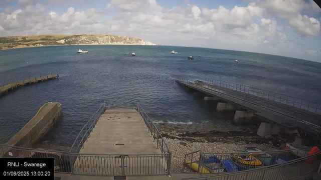 A harbor with a walkway extending into the water, surrounded by a metal fence. Several boats are anchored in the water, with cliffs and a partly cloudy sky in the distance. Near the bottom right, parked small vehicles and possibly kayaks are visible.