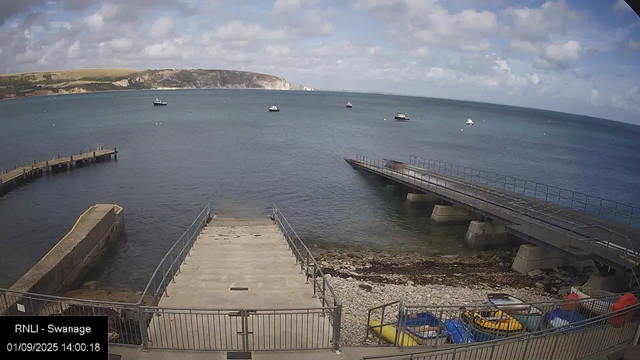 A view of a small harbor with a concrete dock extending into the water, surrounded by a metal railing. Several boats are anchored in the water. The sky is partly cloudy with patches of blue, and distant cliffs can be seen on the horizon. There are some colorful kayaks and small watercraft on the shore near the fenced area.
