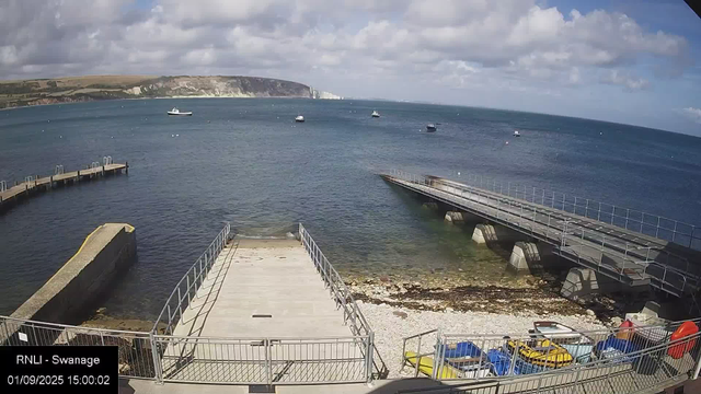 A view of a harbor with a concrete and metal dock extending into the water, surrounded by a metal railing. Several boats and yachts are anchored in the water, and there are parked colorful vehicles on the shore. In the background, a wide bay is visible with a cliff or headland in the distance under a partly cloudy sky.