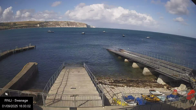 A view of a harbor with two concrete piers extending into the water, one on the left and one on the right, both with metal railings. Several small boats are anchored in the water, with a few near the piers. The water is a deep blue, and there are white clouds in a partly cloudy sky. In the background, a rugged coastline with cliffs and grassy hills stretches across the horizon. In the foreground, there are some colorful kayaks and small boats stored behind a metal fence. The timestamp reads "01/09/2025 16:00:02" and the label indicates this is a webcam from Swansea.