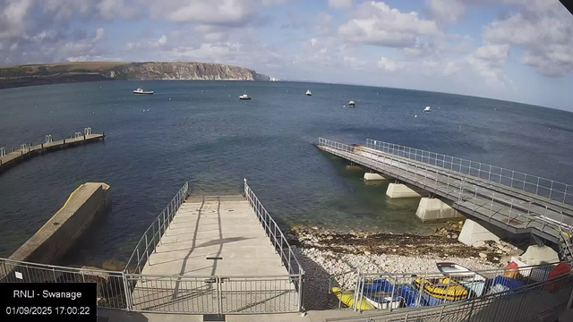 A view of a seaside harbor with several small boats floating on the water, chalk cliffs in the distance under a partly cloudy sky, and a concrete dock extending into the water. There is a metal railing along the dock and some boats and equipment stored near the shoreline.