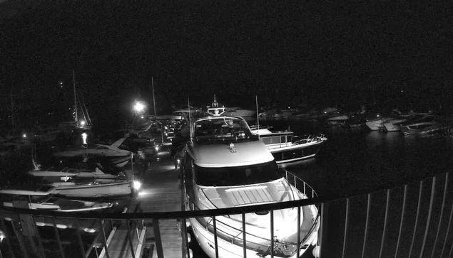 A black and white image of a marina at night showing several boats and yachts docked along the water. There is a person's hand holding a camera or phone slightly above the railing of a balcony or dock in the foreground. The scene is illuminated by a few lights reflecting off the water, creating a quiet and calm nighttime harbor setting.