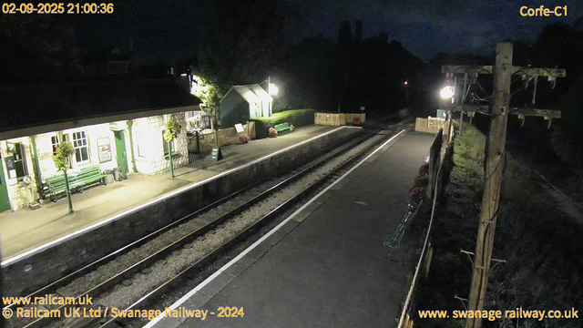 A view of a small railway station platform at night, illuminated by streetlights. There are flower pots and benches along the platform, with a green structure and a small building in the background. The tracks run parallel to the platform, and there are utility poles on the right side of the image. The scene appears quiet with no people visible.
