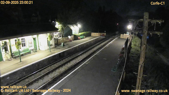 A nighttime view of a small railway station platform illuminated by a few lights. There is a stone building on the left with a green door, windows, and two hanging flower baskets. A green bench and a small green shed are also visible near the building. The railway tracks run between the platform and a fence with fences on the right side, where a wooden electric pole with wires is also seen. The platform appears quiet and empty. The timestamp shows "02-09-2025 23:00:21."