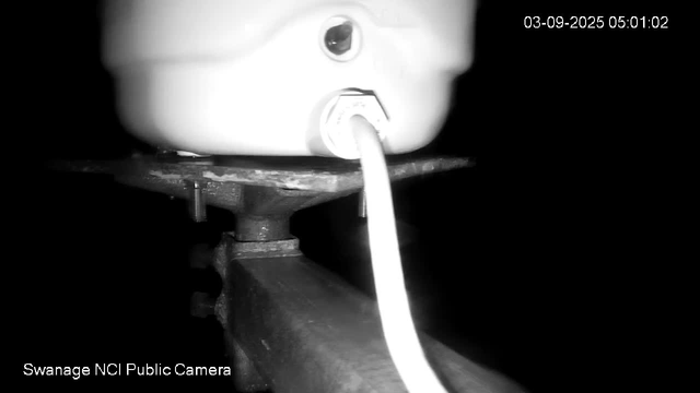 A black-and-white image from a webcam shows a close-up view of an electronic device mounted on a metal bracket. A cable extends from the device downward. The timestamp in the upper right corner reads "03-09-2025 05:01:02". The bottom left corner contains the text "Swanage NCI Public Camera."