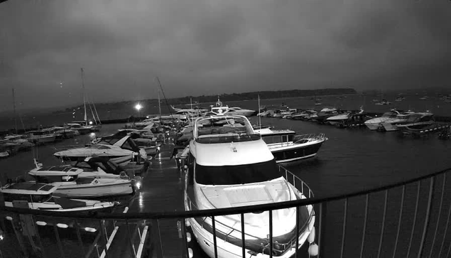A black-and-white image of a marina filled with various boats and yachts docked side by side. The scene is captured from a vantage point behind a curved railing, with numerous vessels and masts visible on a cloudy, overcast day. The water appears calm, and distant land or hills are visible in the background under a dark sky.