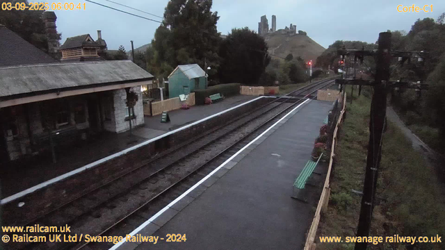 A small train station platform with a building on the left, a green waiting shelter, and flower pots along the fence. Train tracks run through the center, with two red signal lights in the distance. There are green benches on the platform, and a telephone pole with wires on the right. In the background, a hill with ruins of an ancient castle or fortress is visible under a cloudy sky. The timestamp shows the date as March 9, 2025, at 6:00 AM.
