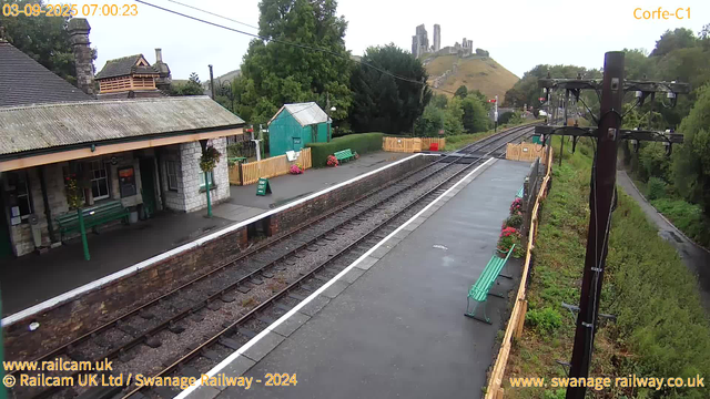 A small railway station platform with a building on the left side featuring a sloped, weathered roof and stone walls. There are green benches and hanging flower baskets on the platform. A sign indicates a "Way Out" entrance. In the background, there is a small teal-colored shed, trees, and a distant hill with ruins or a castle on top. Two railway tracks run parallel along the platform. On the right side, there's a tall wooden utility pole with electrical wires and a narrow green footpath bordered by greenery. The timestamp shows the date as September 3, 2025, at 7:00 AM.