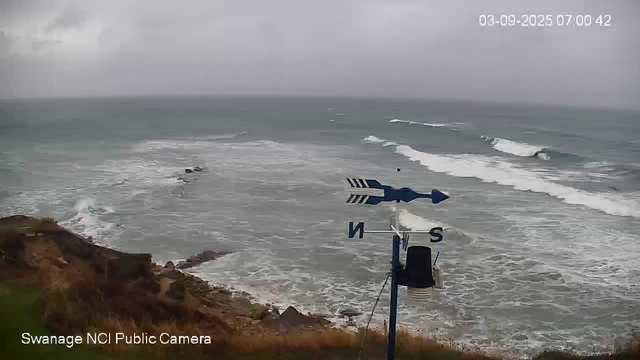 An ocean view from a webcam shows waves crashing onto the shore under a cloudy sky. A weather vane with an arrow and cardinal direction markers (N, S, E, W) is visible in the foreground, mounted on a pole. The timestamp on the image indicates it was taken on September 3, 2025, at 7:00 AM.