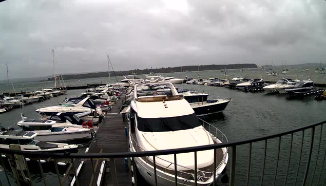 A marina filled with various boats and yachts docked along a wooden pier under a cloudy sky. The scene includes multiple boats lined up on both sides of the pier, with some boats on the water and others moored to the dock. In the background, there is a distant shoreline with more boats floating in the water. The overall atmosphere appears overcast and grey.