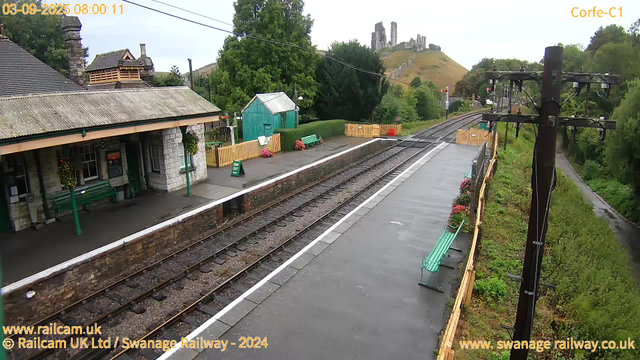 A small train station platform with green benches, a green wooden shed, and a yellow safety fence. There are flower pots with pink and red flowers along the fence. To the right, there are two wooden benches with potted flowers. A lamppost with electrical wires towers over the platform. A railway track runs parallel to the platform, extending into the distance. In the background, there's a hill with an ancient castle ruins on top, surrounded by greenery and trees. The sky is overcast.