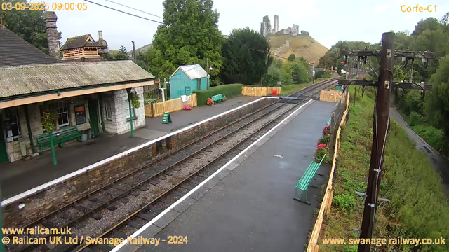 A small railway station platform with a stone building that has a tiled roof and green benches. There are flower pots with pink and red flowers on the platform, along with a sign indicating the exit. The railway tracks run alongside the platform, and there are green hedges and trees in the background. In the distance, a hill with a castle ruin or medieval fortress is visible atop it. To the right, there are wooden utility poles with wires and a narrow pathway surrounded by greenery. The sky is clear, and the scene is well-lit.