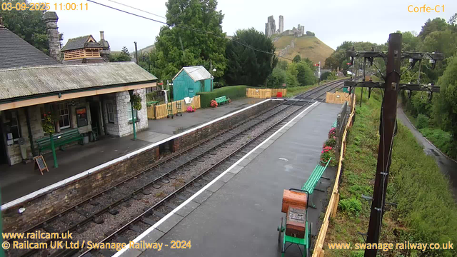A small train station platform with green benches and flower pots, next to railway tracks. There is a stone building with a corrugated roof on the left, with hanging flower baskets near the entrance. In the background, a green shed and a hedge are visible, with colorful flowers near them. A hillside with a castle ruins sits in the distance, surrounded by trees and greenery. A tall wooden telegraph pole with power lines runs along the right side, and a narrow path runs parallel to the tracks behind a green fence. The sky is overcast.