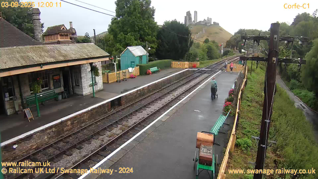 A small train station platform with a stone building on the left, green benches, and colorful flowers along the edge. There are a few people standing and walking on the platform, some holding umbrellas. Railway tracks run through the middle, and a hillside with castle ruins is visible in the background. A large electrical pole with wires extends along the right side of the image. The overall scene appears damp and overcast.