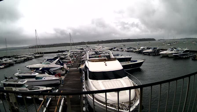 A marina filled with numerous docked boats and yachts under a cloudy sky, with a wooden pier extending into the water and a metal railing in the foreground.