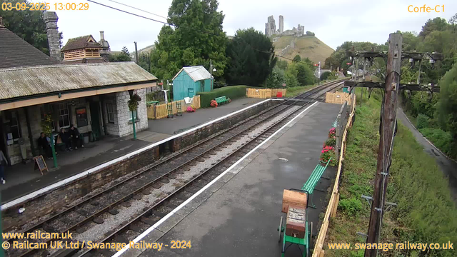 A small train station platform with a stone building on the left, where a few people are sitting. There are colorful flowers in pots and benches along the platform. Rail tracks run through the center, with a green shed and trees behind. In the background, a hill with ruins or a castle is visible, and there are overhead electrical wires and poles along the tracks. The sky is overcast.