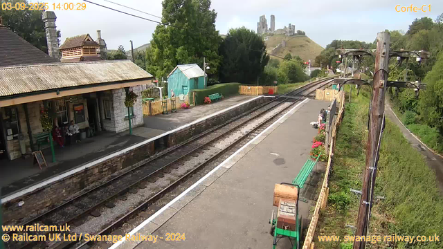 A small railway station with a single platform, two people sitting on a bench under a roof, colorful flower pots along the edge of the platform. The platform is bordered by a yellow fence on the far side, with a small teal-colored building and a green bench nearby. In the background, there are lush green trees, a hill with a castle ruin on top, and a partially cloudy sky. There are electrical poles along the railway tracks, which extend towards the horizon. The date and time are displayed at the top left corner.