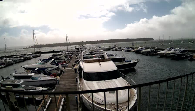 A marina filled with numerous boats and yachts docked at wooden piers, with some boats extending into the water. In the background, there is a distant shoreline with a partly cloudy sky overhead. The photo is taken from a viewpoint behind a black railing.