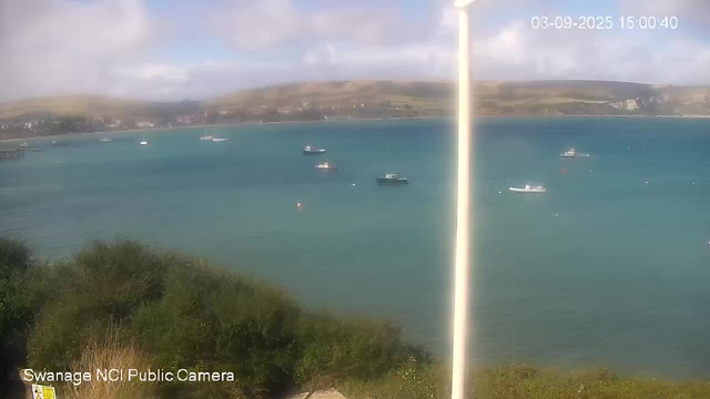 A view of a wide body of water with several boats scattered across the surface. In the foreground, there is green foliage, and in the background, there are hills or cliffs along the shoreline under a partly cloudy sky. The image has a timestamp in the upper right corner reading "03-09-2025 15:00:40" and a label in the lower left corner that says "Swanage NCI Public Camera."