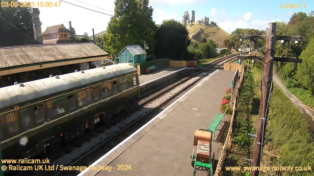 A vintage green train is stopped at a small countryside railway station. The platform has a few green benches, a small turquoise shed, and a row of colorful flower pots along the fence. To the right, a tall wooden utility pole with wires extends upward. In the background, there are trees, rolling hills, and a hilltop castle ruin with stone walls and towers. The sky is partly cloudy with patches of blue. The timestamp shows it is mid-afternoon on September 3, 2025.
