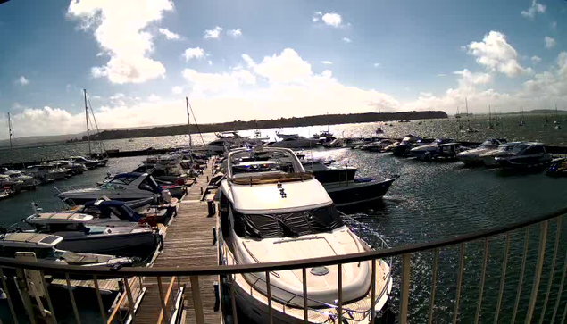 A marina filled with numerous boats and yachts docked along wooden piers, with a clear blue sky and scattered white clouds overhead, and calm water reflecting the sunlight.